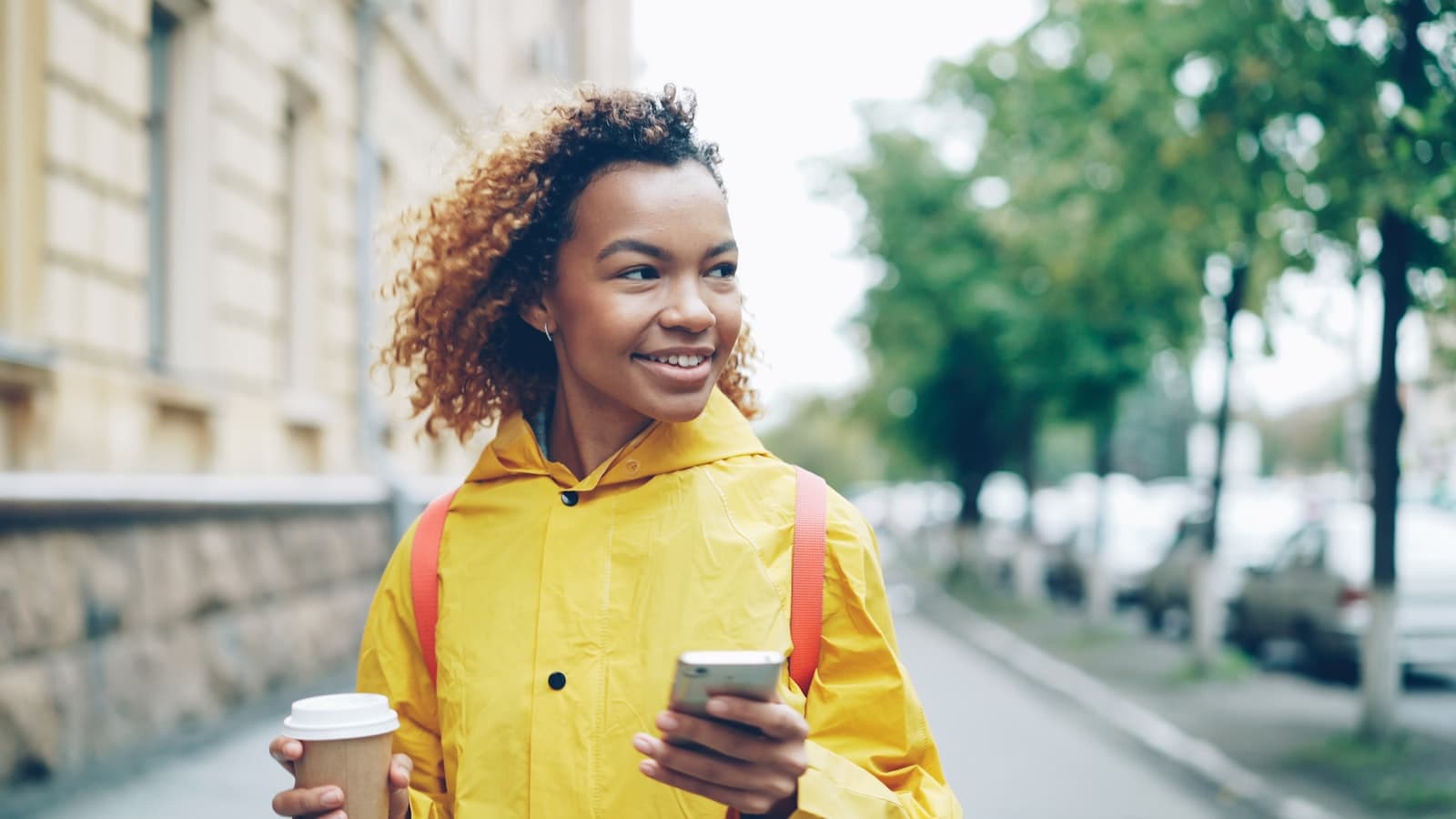 Mulher sorrindo na rua, com um café numa das mãos e o celular na outra